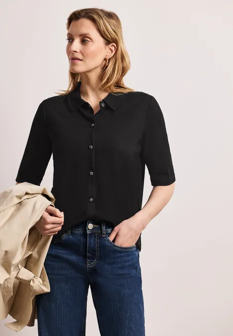 Frau in schwarzem Halbarmhemd und Jeans, hält einen beigen Mantel.
